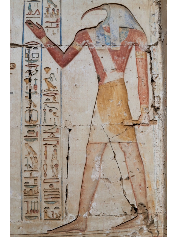 who-is-thoth-ancient-wisdom-egypt Thoth depicted with an ibis head, holding a scroll, standing in front of hieroglyph-covered temple walls.