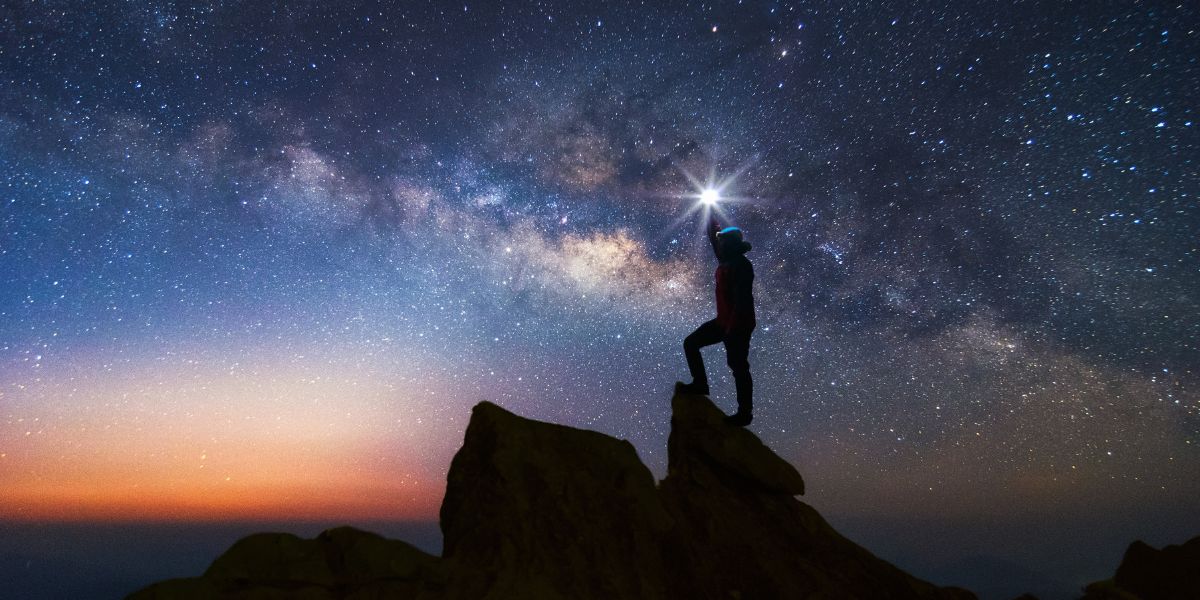 Standing on the Edge of a New Cosmic Dawn Silhouette of a person standing on a cliff under a starry night sky with soft morning light glowing on the left horizon