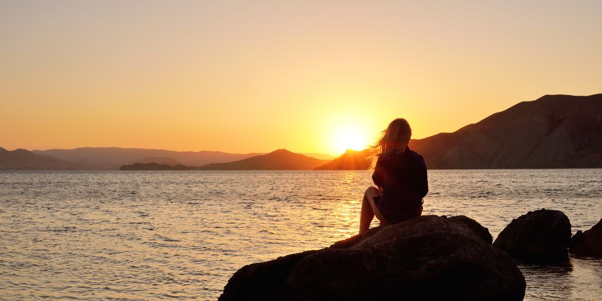 Woman sitting quietly by the water at sunset, reflecting on finding happiness in God